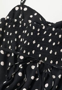 Robe noire à pois blancs, avec un corsage smocké et des bretelles ajustables. Des liens à la taille améliorent le design. Tissu doux et fluide.