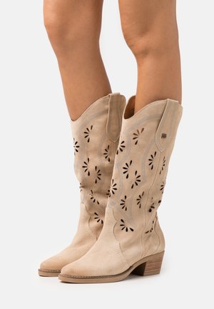 Bottes de cowboy en daim beige avec des motifs décoratifs découpés, des bouts pointus et des talons empilés. Texture lisse avec des accents de couture.