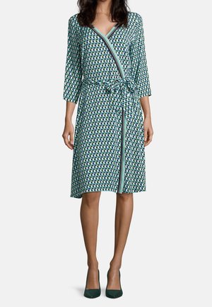 Femme portant une robe portefeuille mi-longue avec des motifs géométriques bleu, vert et blanc, manches trois-quarts, et des talons verts foncés.