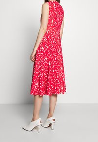Mujer vistiendo un vestido midi rojo sin mangas con manchas abstractas blancas, combinado con unos zapatos de tacón blancos que tienen detalles de lazo en la parte trasera.