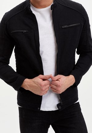 Homme portant une veste noire zippée sur une chemise blanche, les mains tenant la fermeture éclair de la veste près de la taille, sur un fond clair uni.