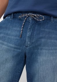 Pantaloni in denim di un blu medio con superficie strutturata, dotati di vita con cordino e dettagli di cucitura discreti sulle tasche.