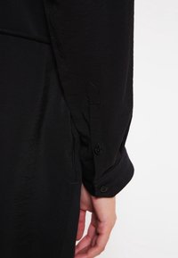 Detalles de una camisa negra de manga larga que muestra un puño abotonado y una textura suave, con un aspecto ligeramente arrugado y un ajuste entallado.