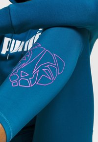 Tyrkysové sportovní legíny vyrobené z elastického materiálu; na levém koleni se nachází růžová geometrická grafika lva a logo na oblasti boku.