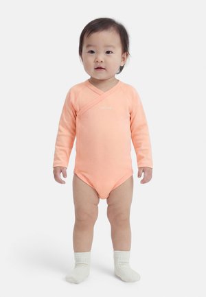 Νήπιο όρθιο, φοράει ένα μακρυμάνικο ροδακινί φορμάκι και λευκές κάλτσες, σε απλό λευκό φόντο.