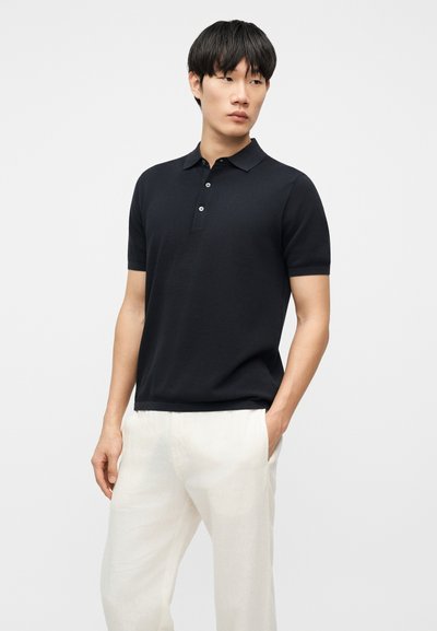 Άνδρας που φοράει μαύρο πόλο με κοντά μανίκια και λευκό παντελόνι, στέκεται με το ένα χέρι στην τσέπη του, κοιτάζοντας προς τα πλάγια.