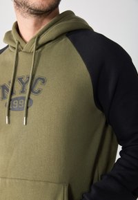 Hoodie vert olive avec des manches raglan noires, présentant une poche kangourou à l'avant et un graphisme NYC, complété par un cordon de serrage et des embouts en métal.