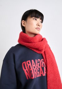 Bufanda roja y peluda de material suave, drapeada alrededor del cuello, combinada con una sudadera azul marino que presenta un texto rojo audaz.