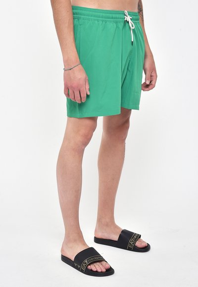 Pantaloni da bagno verdi con elastico in vita e lacci bianchi. Abbinati a sandali neri a infradito con brandizzazione dorata.