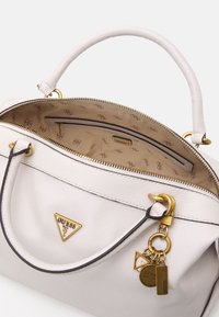 Biała skórzana torebka Guess z złotym logo, z zamkiem otwartym, ukazującym beżowe wnętrze z wzorem logo, z dołączonymi złotymi zawieszkami w kształcie breloczków.