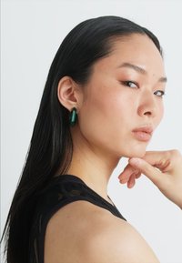 Green cylindrical earrings displayed on a model with long dark hair, wearing a black textured top. Smooth finish with a sleek design.
