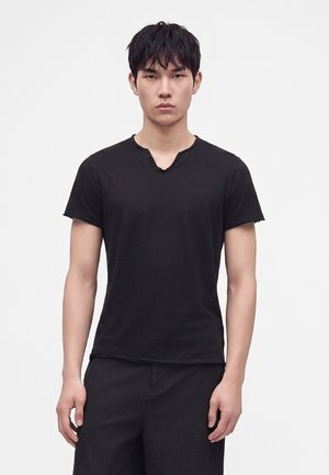 Jeune homme aux cheveux noirs courts portant un t-shirt noir uni à manches courtes et un pantalon noir sur fond blanc.