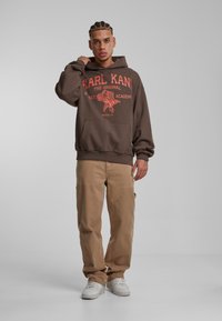 Sudadera marrón con texto gráfico rojo y un diseño de pájaro, con un bolsillo delantero y puños acanalados. Combinada con pantalones cargo beige y zapatillas blancas.