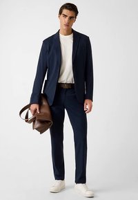 Veste de costume bleu marine portée sur un t-shirt blanc, associée à un pantalon bleu marine ajusté et des baskets blanches. Sac en cuir marron au design structuré.