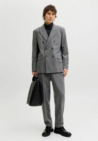 Grå dobbeltspent dress laget av teksturert stoff, kombinert med en tettsittende svart høyhalset genser og svarte skinnsko. En svart totebag holdes i hånden.