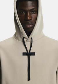 Sweatshirt à capuche en tissu beige clair avec une coupe décontractée, arborant des cordons noirs et une étiquette carrée indiquant "INGÉNIERIE SUISSE."