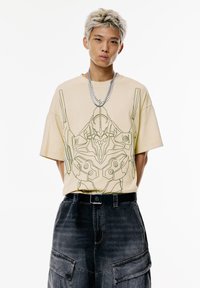 Beige, oversized póló zöld grafikával robot részletekkel. Fekete cargo sorttal és fekete övvel viselve. Ezüst láncnyaklánc.