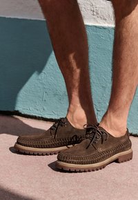 Chaussures en daim marron avec un design tissé, fermeture à lacets et semelle épaisse en caoutchouc. Style bas laissant apparaître la cheville, sur un fond coloré.