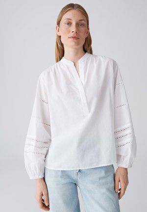 Femme aux cheveux blonds raides portant un chemisier blanc avec de la dentelle œil-de-perdrix sur les manches bouffantes et un jean bleu clair, debout devant un fond uni.