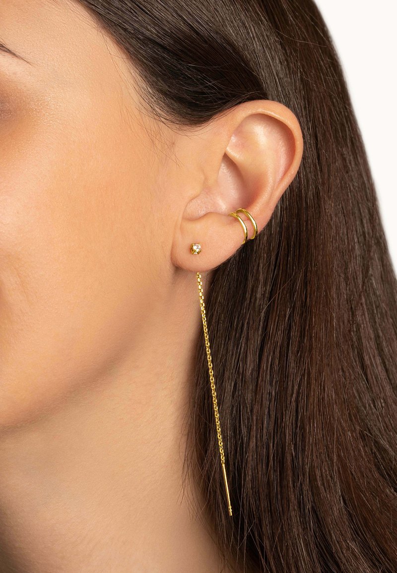 Goldene Creolen mit einem kleinen Edelstein am Ohrläppchen und einem hängenden Kettenakzent. Die Kette hat am Ende einen dünnen Stab.