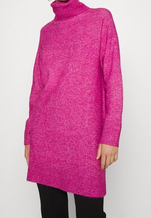 Kobieta ubrana w jaskrawo różową dzianinową sukienkę z długim rękawem i golfem, z prążkowanymi mankietami i brzegiem, w połączeniu z czarnymi spodniami.