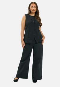 Mujer con cabello largo que lleva una blusa sin mangas negra de botones, pantalones negros de pierna ancha y zapatos negros, de pie con una mano en la cadera.