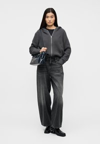 Γυναίκα που φοράει μαύρο γκρι ζακέτα με φερμουάρ, μαύρο ξεθωριασμένο τζιν με φαρδιά παντελόνια, μαύρες μπότες και κρατάει μια σκούρα μπλε τσάντα ώμου.