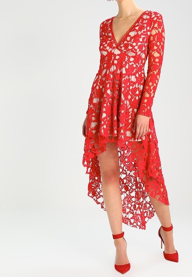 Robe en dentelle rouge à manches longues et décolleté plongeant. Ourlet asymétrique, texture transparente, motif floral complexe, associée à des talons rouges.