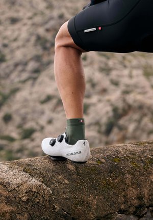 Weiße Radschuhe mit schwarzen Schnallen, strukturiertem Obermaterial und Specialized-Logo. Olivgrüne Socke mit kleinem rotem Emblem. Bein einer Person auf einer steinernen Oberfläche.