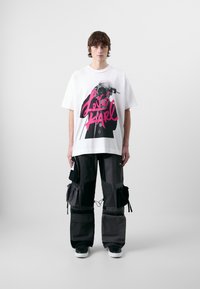 Witte katoenen t-shirt met grote roze grafische tekst "Love Karl" en een zwarte illustratie. Het model draagt cargo-stijl zwarte en grijze broek.