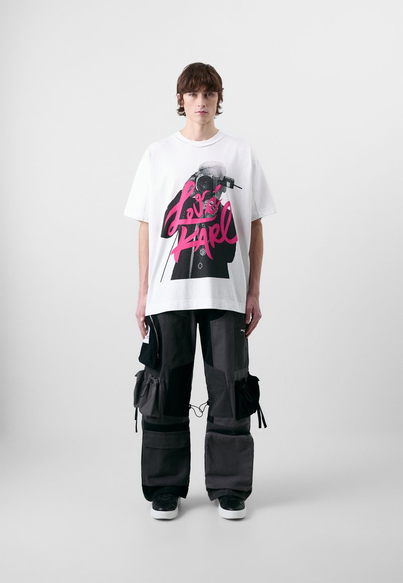 Witte katoenen t-shirt met grote roze grafische tekst "Love Karl" en een zwarte illustratie. Het model draagt cargo-stijl zwarte en grijze broek.