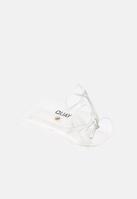 Des lunettes de chat claires reposant dans un étui transparent avec fermeture à bouton doré ; dotées d'un design minimaliste et du nom de la marque "QUAY" imprimé.