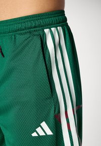 Calções atléticos verdes com um tecido texturizado, riscas brancas verticais e um logótipo em bordô. Apresentam uma cintura elástica e um bolso lateral.