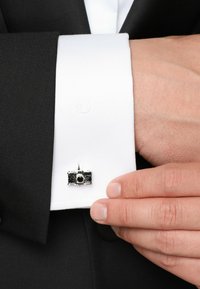 Srebrny spink w kształcie aparatu przymocowany do białej koszuli, z widoczną czarną marynarką. Teksturowany materiał na rękawie.