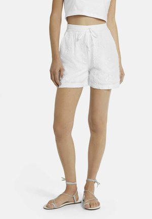 Frau trägt weiße bestickte Shorts mit elastischem Bund und Kordelzug, kombiniert mit hellgrauen, flachen Sandalen mit Riemchen.