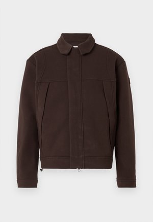 Braune Jacke aus weichem Material mit einem Kragen, vorderem Reißverschluss und zwei seitlichen Taschen. Sie zeichnet sich durch minimale Nähte und ein schlichtes Design aus.