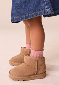 Bottes en daim marron clair à la cheville avec une finition texturée, associées à des chaussettes roses côtelées, vues de profil sur un fond doux et neutre.