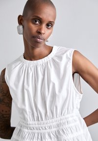 Rochie albă fără mâneci cu decolteu pliat și talie elastică. Material neted, formă structurată și strălucire subtilă. Cercei mari cu textură în formă de pătrat.