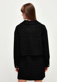 Schwarze Tweedjacke mit einem cropped Design, breitem Kragen und geknöpften Manschetten. Getragen über einem passenden schwarzen Rock mit einer strukturierten Oberfläche.