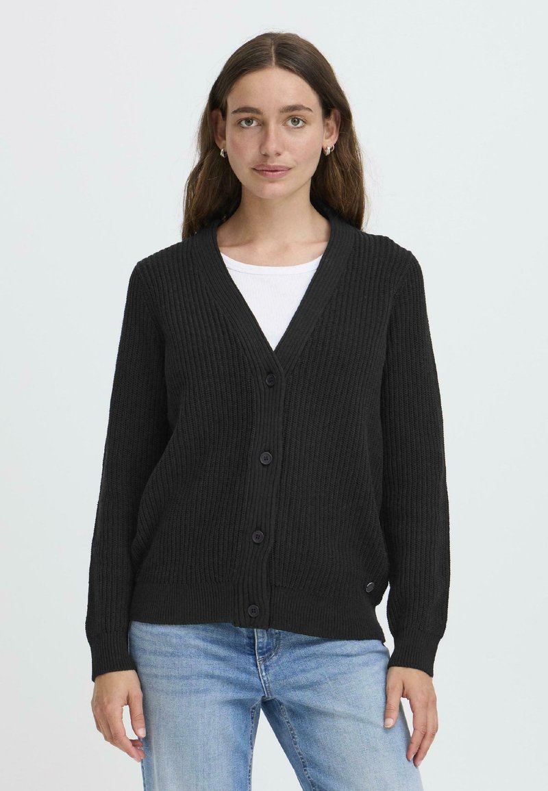 Cardigan noir côtelé avec un col en V, doté de six boutons et de manches longues. Porté par-dessus une chemise blanche, à la texture lisse et à la coupe décontractée.