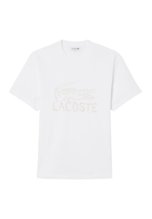 T-shirt bianca a maniche corte con scollo rotondo, logo coccodrillo Lacoste in rilievo e scritta "LACOSTE" al centro davanti.