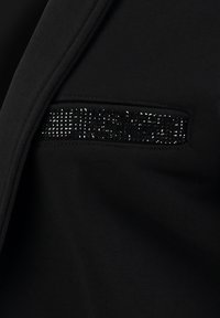 Black garment close-up featuring a textured fabric. A pocket with a row of shiny black rhinestones adds detail. Smooth appearance overall.