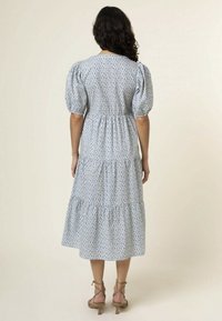 Maxi dress in light blue with a diamond pattern, puff sleeves, fitted waist, and tiered skirt. Flat sandals with ankle straps.