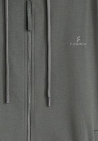 Sudadera con capucha verde oscuro con cremallera, de textura suave, con una cremallera plateada y cordones, y un pequeño logo impreso en el pecho.