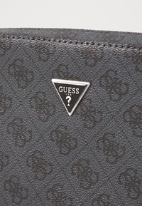 Material negro texturizado con patrón en relieve; presenta una etiqueta triangular plateada con "GUESS" y un signo de interrogación en el interior.