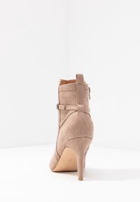 Bottine en daim beige avec un bout pointu, un talon fin, des détails perforés décoratifs et des accents de sangle à l'arrière.