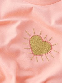 Rožnata tkanina z vezilom v obliki srca v zlati barvi, obdana s sončnimi žarki. Mehka tekstura, srednje težka tkanina in enostavni oblikovalski detajli.