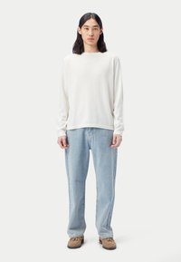 Langärmeliges, elfenbeinfarbenes Hemd aus leichtem Stoff, kombiniert mit hellblauen Jeans. Das Modell steht vor neutralem Hintergrund und trägt braune Schuhe.