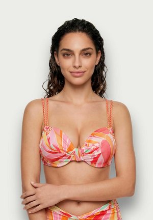 Donna con capelli ricci scuri indossa un top bikini con fantasia rosa, arancione e rossa, in piedi con le braccia incrociate su uno sfondo chiaro e uniforme.