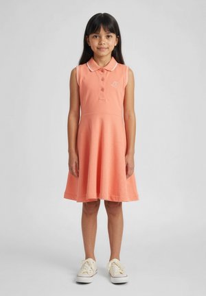 Ragazza giovane in piedi, rivolta verso l'avanti, che indossa un vestito senza maniche color pesca con colletto e scarpe da ginnastica bianche su uno sfondo semplice.